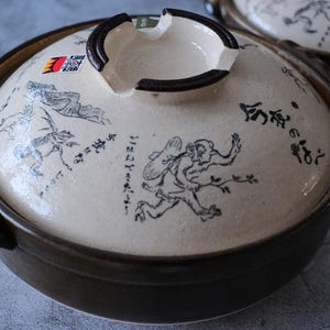 Puede incluir: Olla de cocina japonesa de cer&aacute;mica con tapa. La tapa blanquecina presenta ilustraciones negras y texto japon&eacute;s. La olla tiene un borde y asas de color marr&oacute;n oscuro. Se ve otra olla en el fondo.