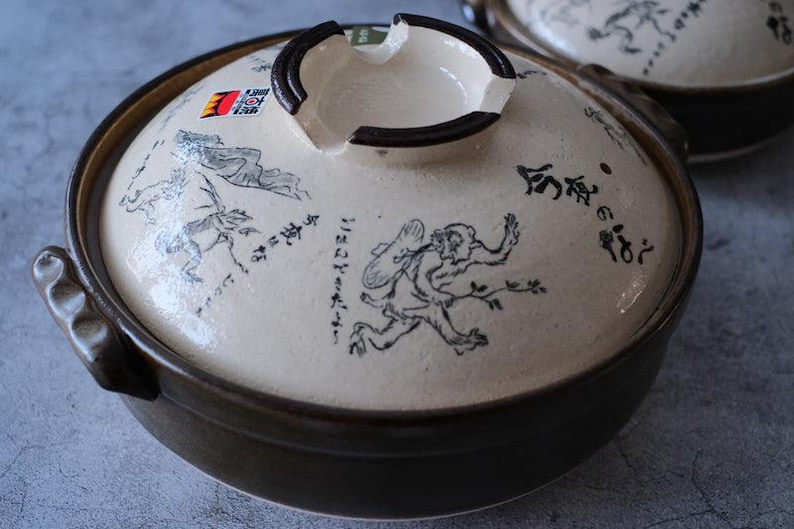 Puede incluir: Olla de cocina japonesa de cer&aacute;mica con tapa. La tapa blanquecina presenta ilustraciones negras y texto japon&eacute;s. La olla tiene un borde y asas de color marr&oacute;n oscuro. Se ve otra olla en el fondo.