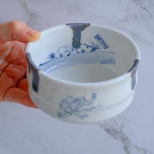 May include: White ceramic bowl with blue painted accents and a stylized elephant design. The bowl has a rounded shape with a slightly flared rim and two small handles. The interior features blue cloud and landscape motifs.