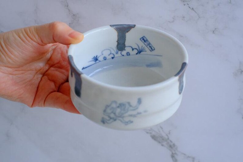 May include: White ceramic bowl with blue painted accents and a stylized elephant design. The bowl has a rounded shape with a slightly flared rim and two small handles. The interior features blue cloud and landscape motifs.