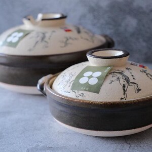 Puede incluir: Dos ollas de cer&aacute;mica de estilo japon&eacute;s con tapas. Las ollas son de color marr&oacute;n oscuro con tapas blanquecinas. Las tapas presentan una ilustraci&oacute;n de rana y una etiqueta verde con c&iacute;rculos blancos. Asas en los laterales.