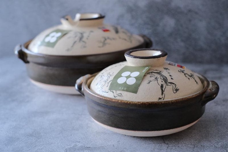Puede incluir: Dos ollas de cer&aacute;mica de estilo japon&eacute;s con tapas. Las ollas son de color marr&oacute;n oscuro con tapas blanquecinas. Las tapas presentan una ilustraci&oacute;n de rana y una etiqueta verde con c&iacute;rculos blancos. Asas en los laterales.