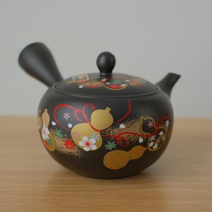 May include: A black ceramic teapot with a rounded body, a curved handle, and a small spout. The teapot is decorated with gold, red, and white floral and gourd designs. The lid has a small black knob.