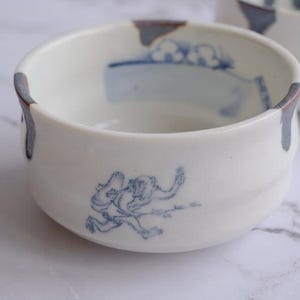 May include: White ceramic bowl with blue illustrations of a figure and landscape elements. The rim has a dark blue and brown accent. A second bowl is partially visible in the background.