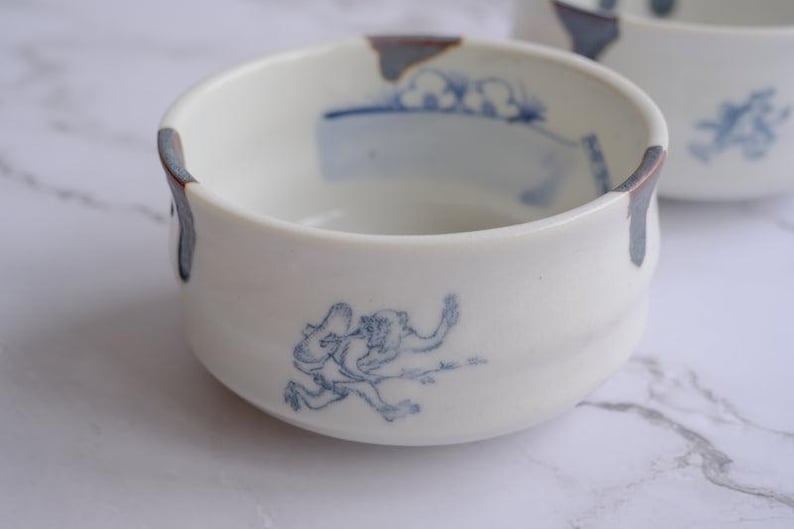 May include: White ceramic bowl with blue illustrations of a figure and landscape elements. The rim has a dark blue and brown accent. A second bowl is partially visible in the background.