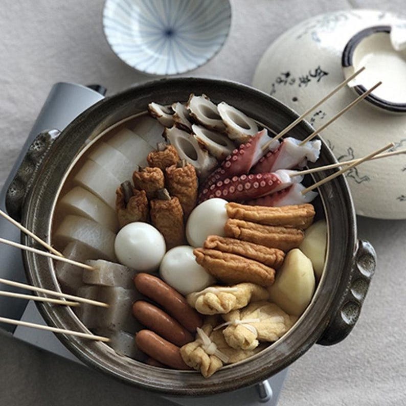 Puede incluir: Primer plano de un plato de oden japon&eacute;s, con una variedad de ingredientes como huevos cocidos, r&aacute;bano daikon, pasteles de pescado, pulpo y salchichas. Los ingredientes est&aacute;n dispuestos en una olla de cer&aacute;mica oscura con brochetas de madera. Un cuenco peque&ntilde;o y una olla de cer&aacute;mica est&aacute;n en el fondo.