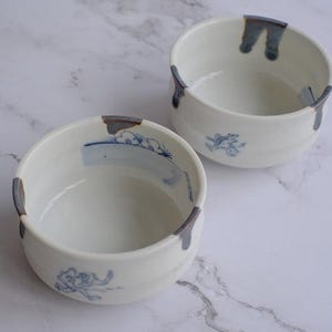 May include: Two white ceramic bowls with blue and gray accents. Each bowl features a hand-painted blue design and a gray accent on the rim. The bowls are round and have a simple, elegant design, perfect for serving food.