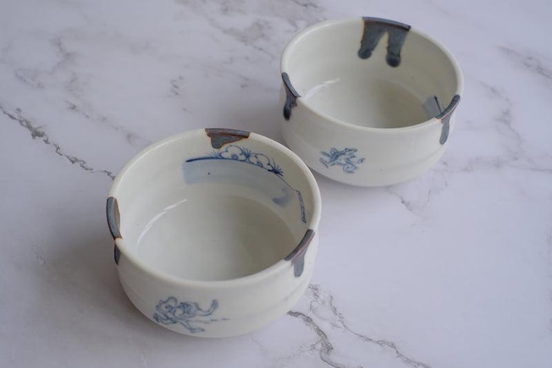 May include: Two white ceramic bowls with blue and gray accents. Each bowl features a hand-painted blue design and a gray accent on the rim. The bowls are round and have a simple, elegant design, perfect for serving food.