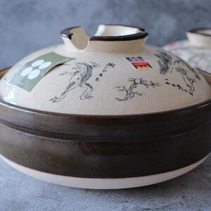 Puede incluir: Una olla donabe japonesa de cer&aacute;mica con tapa. La olla es marr&oacute;n con una tapa color crema decorada con una ilustraci&oacute;n de rana y texto japon&eacute;s. La olla tiene dos asas y una peque&ntilde;a abertura en la tapa.