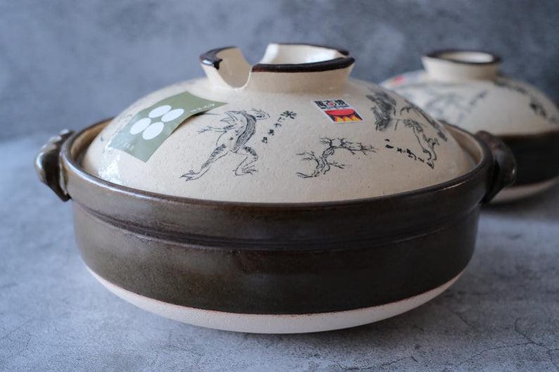 Puede incluir: Una olla donabe japonesa de cer&aacute;mica con tapa. La olla es marr&oacute;n con una tapa color crema decorada con una ilustraci&oacute;n de rana y texto japon&eacute;s. La olla tiene dos asas y una peque&ntilde;a abertura en la tapa.