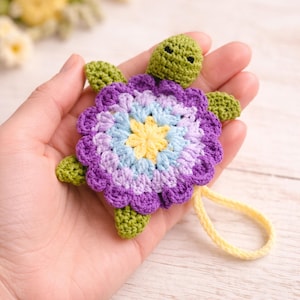 May include: A handmade crochet turtle with a flower-shaped shell. The shell features a gradient of purple, blue, and yellow. The turtle has green head, feet, and a yellow loop attached. The craft is held in a hand.