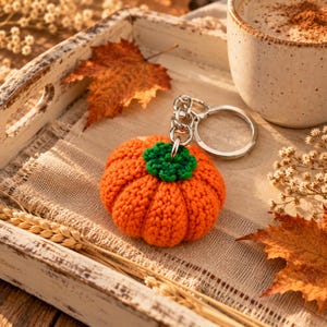 May include: A small, round, orange crocheted pumpkin keychain with a green crocheted stem attached to a silver-toned metal split ring. The pumpkin features a textured, ribbed yarn surface and is crafted with a dense, uniform stitch pattern throughout.