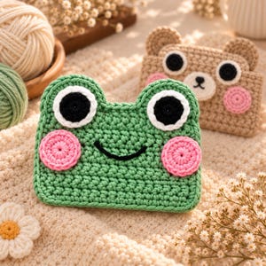 Peut inclure: Une pochette en crochet vert représentant une grenouille avec de grands yeux blancs et noirs et des joues roses circulaires. Derrière se trouve une pochette en crochet beige représentant un ours avec des détails similaires aux yeux et aux joues.