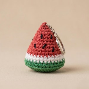 Puede incluir: Un llavero de ganchillo con forma de cuña de sandía que presenta una parte superior triangular roja con semillas bordadas en negro, una banda central blanca y una base verde. Incluye una anilla de metal plateado en la parte superior.