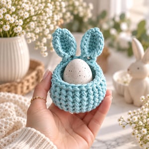 May include: A light blue crocheted egg holder featuring two upright bunny ears and a ribbed texture. The holder contains a single white egg with small black speckles throughout its surface. The item is held in a palm, showcasing its compact size.