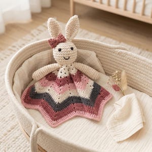 May include: A crochet bunny lovey featuring a cream-colored head with long ears, a small dusty rose bow, and black embroidered eyes and mouth. The attached blanket body is star-shaped with alternating stripes of cream, dusty rose, and dark gray yarn.