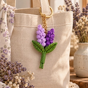 May include: A crochet keychain featuring two lavender flower stalks in light and dark purple yarn, accompanied by two green leaves and a wrapped green stem. The floral charm is attached to a gold-toned metal split ring by a short chain.
