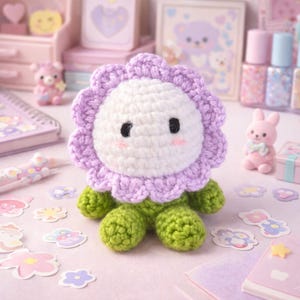 May include: A handmade crochet flower doll with a white center, lavender petals, and green stem and feet. The doll has black eyes and pink cheeks. The doll is surrounded by stickers, a pink bunny figurine, and other decorative items.