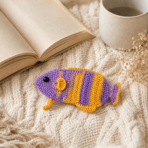 Peut inclure: Un petit poisson plat au crochet, orné de rayures verticales alternées en fil jaune vif et violet clair. Le poisson possède un œil en perle noire, une petite nageoire latérale arrondie et une queue en éventail, le tout finement travaillé.