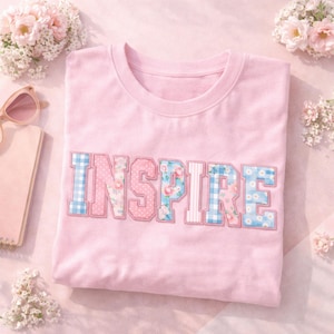 May include: A pale pink t-shirt featuring the word "INSPIRE" in appliqued letters. The letters use floral, polka dot, and gingham patterns in pink and blue. The shirt is neatly folded on a pink surface, with floral accents.