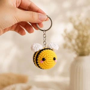 Puede incluir: Un llavero de abeja tejido a ganchillo de forma redonda con un cuerpo de rayas amarillas y negras, dos pequeñas alas blancas, ojos de cuentas negras y diminutas mejillas rosadas bordadas. La abeja está unida a una anilla de metal plateado.