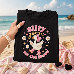 Pode incluir: Uma t-shirt preta com um gráfico de um ganso branco a usar galochas cor-de-rosa às bolinhas e um laço cor-de-rosa. O texto "silly goose on the loose" está escrito em letras cor-de-rosa e amarelas, rodeado por margaridas brancas, corações cor-de-rosa e estrelas amarelas.