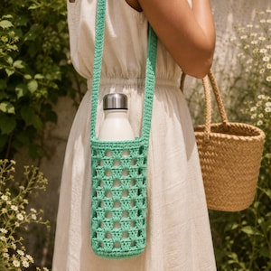 Könnte beinhalten: Ein mintgrüner gehäkelter Wasserflaschenhalter mit langem Schulterriemen und einem durchbrochenen Granny-Square-Muster. Der Halter enthält eine weiße Wasserflasche mit einem silberfarbenen Metalldeckel zum Transport von Getränken.