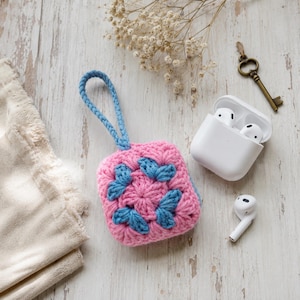 Pode incluir: Uma capa quadrada de crochê para fones de ouvido sem fio com um padrão floral rosa e azul e uma alça azul correspondente. A capa é acompanhada por um estojo de carregamento branco com dois fones, um dos quais fora do estojo aberto.