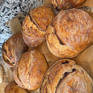 May include: Overhead shot of several freshly baked loaves of sourdough bread. The crusts are golden brown with decorative scoring. Some loaves have visible inclusions. The bread rests on brown parchment paper.