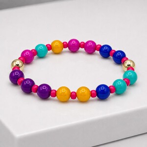 May include: A circular beaded bracelet featuring a repeating pattern of glossy spherical beads in shades of magenta, turquoise, yellow, purple, and blue, separated by small hot pink spacer beads and two metallic gold-toned spherical beads.