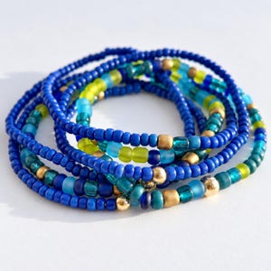 May include: A stack of beaded bracelets featuring a mix of small round beads in shades of royal blue, turquoise, and lime green. The design incorporates metallic gold-toned spherical beads interspersed throughout the blue and green glass beads.