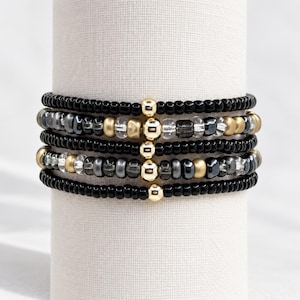 May include: A stack of five beaded bracelets featuring small black beads, metallic gold-tone spheres, clear faceted beads, and dark grey metallic beads. Each bracelet is accented with a single central gold-tone bead aligned vertically.
