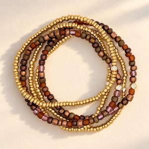 May include: A set of three circular bracelets composed of small beads in metallic gold, matte brown, and translucent mauve. The beads are arranged in alternating patterns, with some sections featuring small, round, gold-toned metal spacers.