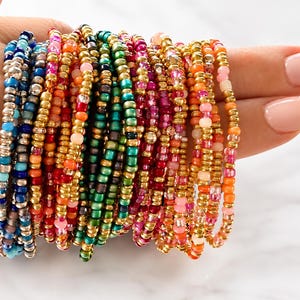 May include: A collection of multiple elastic bracelets made from small, round glass beads in a variety of colors including blue, green, pink, orange, and red, interspersed with metallic gold and silver beads arranged in a dense, stacked formation.