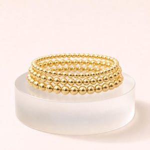 May include: A set of three stacked bracelets featuring polished gold-tone spherical beads of varying sizes. The bracelets are arranged in a circular formation, showcasing a reflective metallic finish and a uniform, smooth, round bead design throughout.