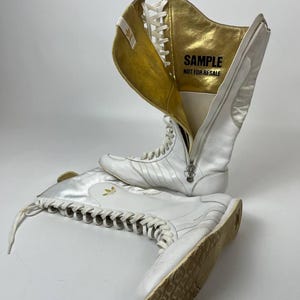 Puede incluir: Zapatillas altas blancas con detalles dorados y cierre de cremallera. Las zapatillas tienen una etiqueta de muestra en el interior y el logo de Adidas. Los zapatos están atados con cordones y tienen un diseño retro.