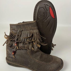 May include: Brown leather ankle boots with fringe detailing and a zipper closure. The boots feature a decorative beaded accent and the brand name "Kickers" is visible. The sole of the boot has a cartoon graphic.
