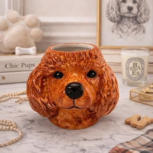 May include: A ceramic dog-shaped container with a brown, textured exterior and black nose and eyes. The container is designed to resemble a poodle. The container is sitting on a marble surface. The words "Chic Pups" are visible on a book.
