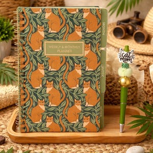 May include: A spiral-bound weekly and monthly planner with a cat pattern. The cover features orange cats on a dark green background. A green pen with a cat mom charm and a wooden tray are also in the image.
