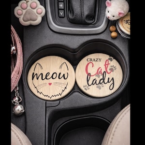 May include: Two round car coasters with cat-themed designs. One coaster says "meow" with a cat face, and the other says "Crazy Cat Lady" with paw prints. The coasters are light brown with black and red text.