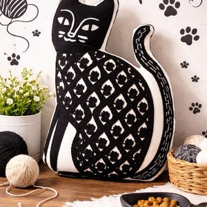 May include: A black and white cat-shaped pillow with a decorative pattern. The pillow is on a wooden surface with yarn balls, a small plant, and a basket. The background has paw prints and cat-themed drawings.