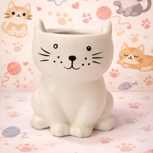 May include: A white ceramic cat-shaped planter with a smiling face, whiskers, and small ears. The planter is sitting on a pink background with cat paw prints and cat illustrations. This decorative planter is ideal for small plants.
