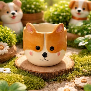 May include: A ceramic planter shaped like a corgi dog's face, with orange and white coloring, black eyes, and pink cheeks. The planter sits on a wooden slice surrounded by greenery and other corgi-themed planters.