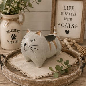 May include: A ceramic cat-shaped planter with a glossy white finish and tan stripes. The cat has closed eyes, a black nose, and whiskers. It is positioned on a square piece of beige fabric inside a round wooden tray with metal handles.
