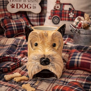 May include: A ceramic dog-shaped container with a tan and black color scheme. The container is surrounded by dog-themed accessories, including a plaid pillow, a dog collar, and bone-shaped dog treats. The sign says "Home is where the DOG is."
