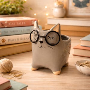 May include: A gray ceramic cat-shaped planter with black-rimmed glasses and closed eyes. The planter has a small opening at the top and is sitting on a wooden surface. Books with titles like "Cat Tales" and "Paws & Relax" are in the background.