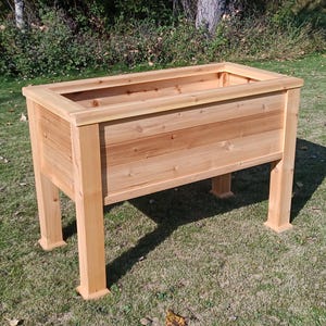 May include: A rectangular, raised wooden planter box with a natural finish. The planter has four sturdy legs and is constructed from horizontal wooden planks. The box is empty and sits on a grassy surface, ideal for gardening.