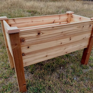 May include: A rectangular wooden planter box with a natural finish. The planter has a slatted design and is supported by four sturdy legs. The corners are reinforced with decorative caps. This outdoor planter is ideal for gardening.