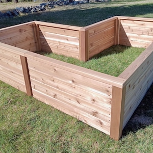 May include: A large, L-shaped wooden raised garden bed made of light-colored wood. The bed is constructed with horizontal planks and vertical supports, sitting on a grassy lawn. The bed is approximately 60 inches wide and 60 inches long.
