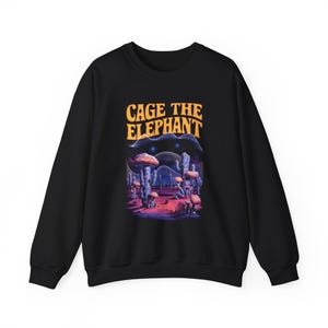 May include: Black crewneck sweatshirt featuring the text "CAGE THE ELEPHANT" in orange above a colorful graphic. The graphic depicts a desert landscape with mushrooms and cacti under a starry sky. A stylish and comfortable apparel item.